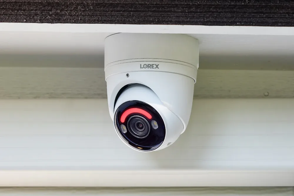 White security camera with a glowing red ring, labeled "LOREX," mounted under a roof with wood texture and a light background.